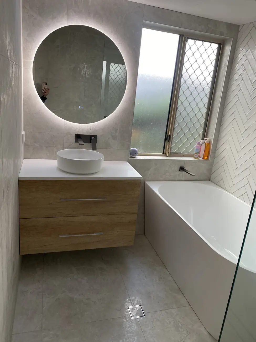 Bathroom with a backlit round mirror above a timber vanity and a bath beside the window